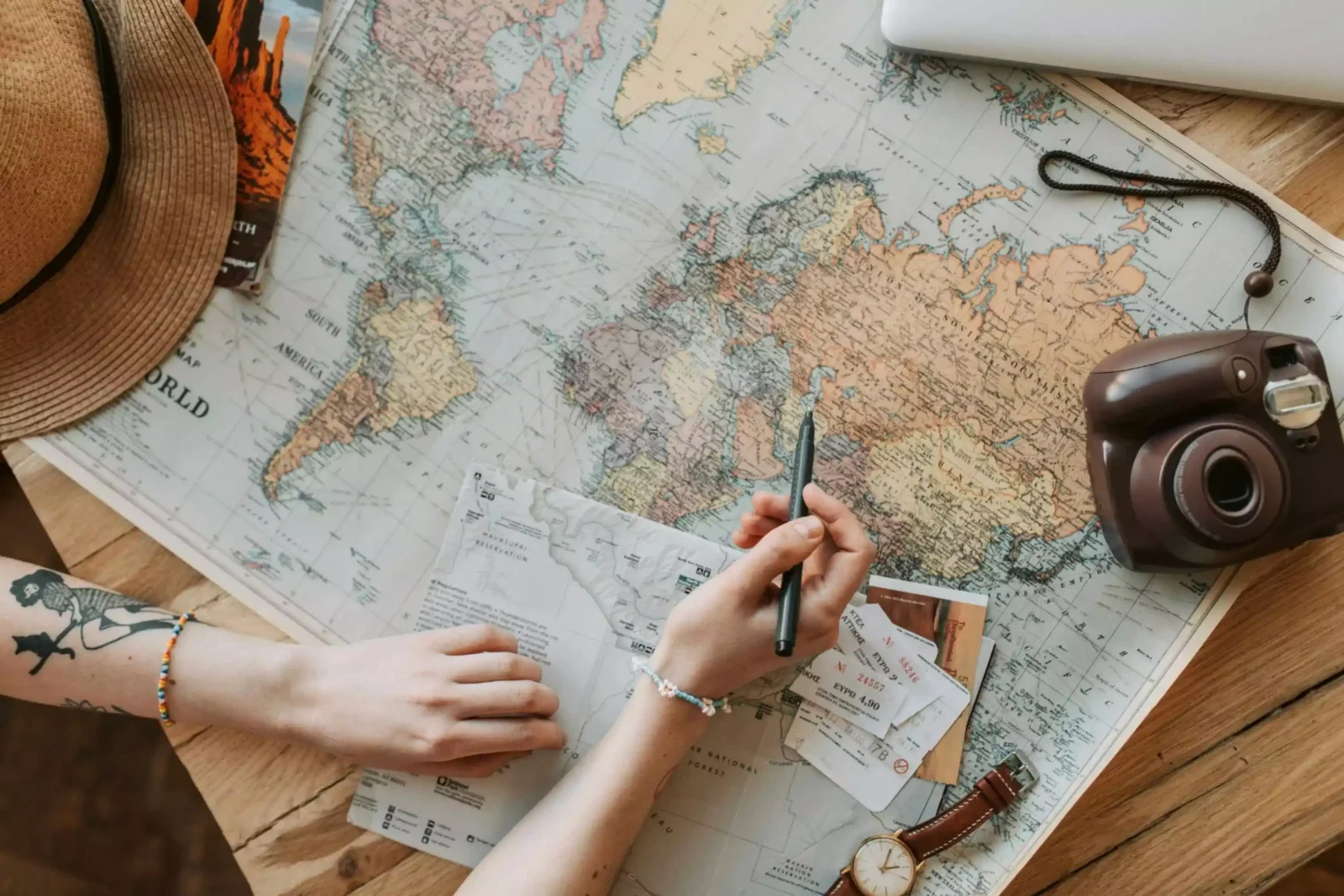 A top-down view of a traveler planning a trip on a wooden desk, featuring a vintage-style world map, a camera, a travel hat, and a hand holding a pen over Europe, symbolizing the ease of choosing the best eSIM for international travel in 2026.