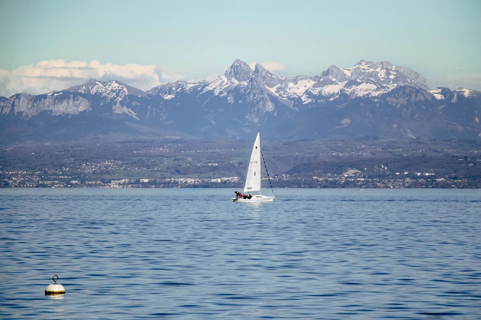 A sailboat on Lake Geneva facing the snow-capped peaks of the Swiss Alps under a clear sky