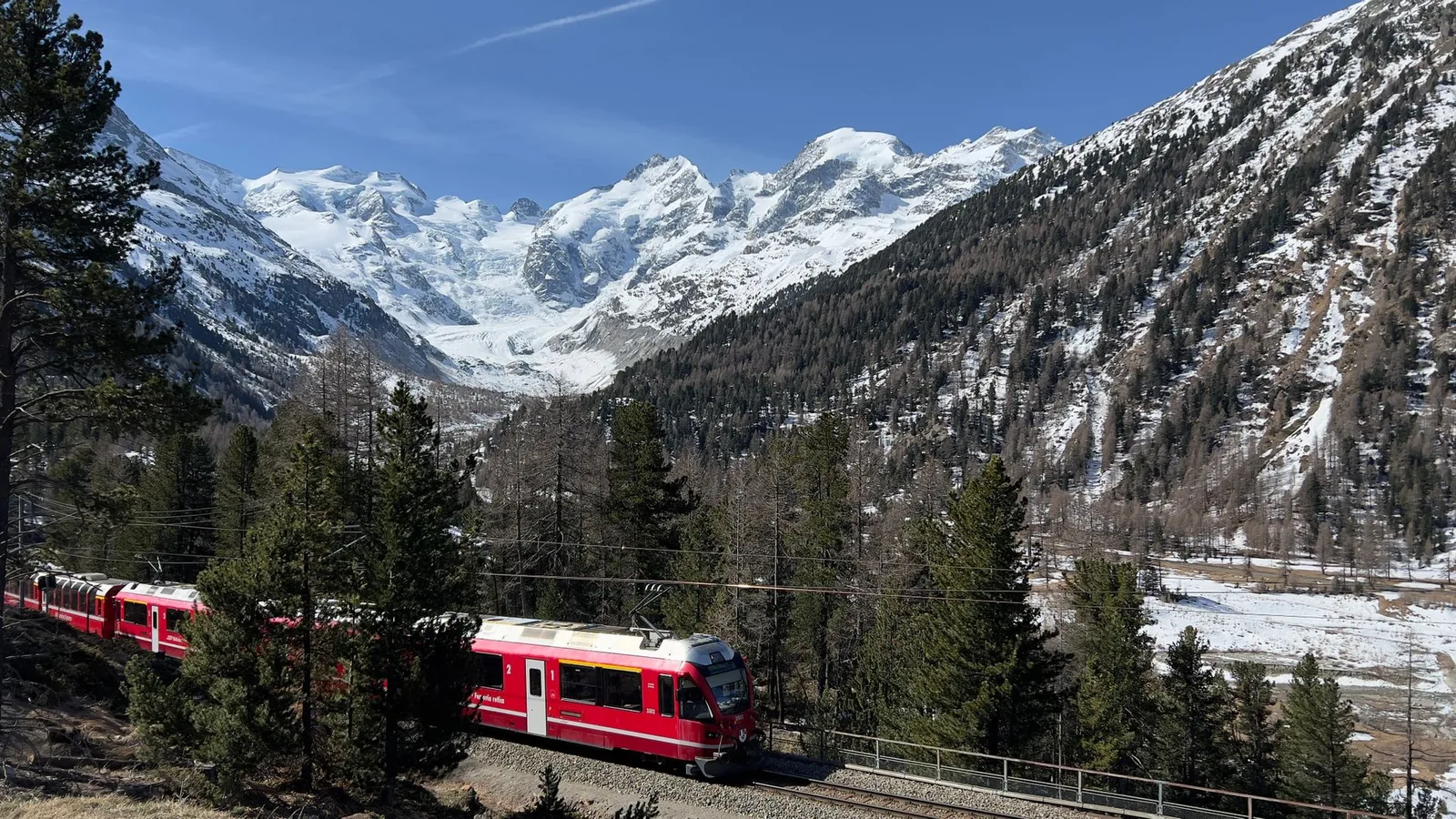 A red Bernina Express train crossing a valley facing the snow-capped glaciers of the Swiss Alps