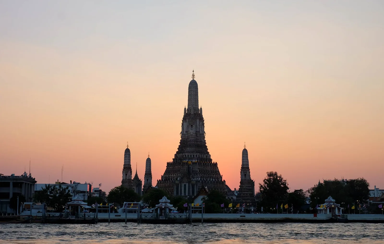 The Wat Arun temple on the banks of the Chao Phraya River in Bangkok at sunset, its spire silhouetted against an orange sky