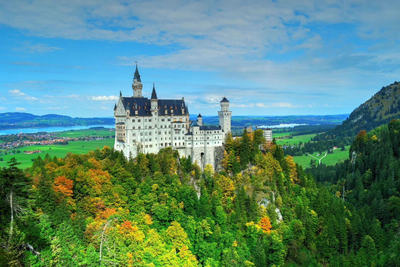 Neuschwanstein Castle perched on its wooded hill in autumn colours, with the Bavarian Alps and a lake in the background