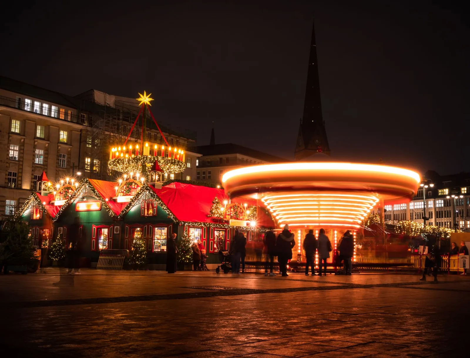 German Christmas market at night with illuminated wooden chalets, a golden carousel and a church spire behind